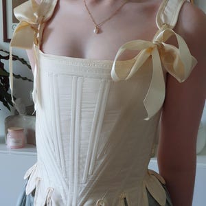 May include: Cream-colored corset with vertical stitching and decorative scalloped hem. The corset features wide shoulder straps tied with large cream-colored bows. A gold necklace with a pearl pendant is also visible.