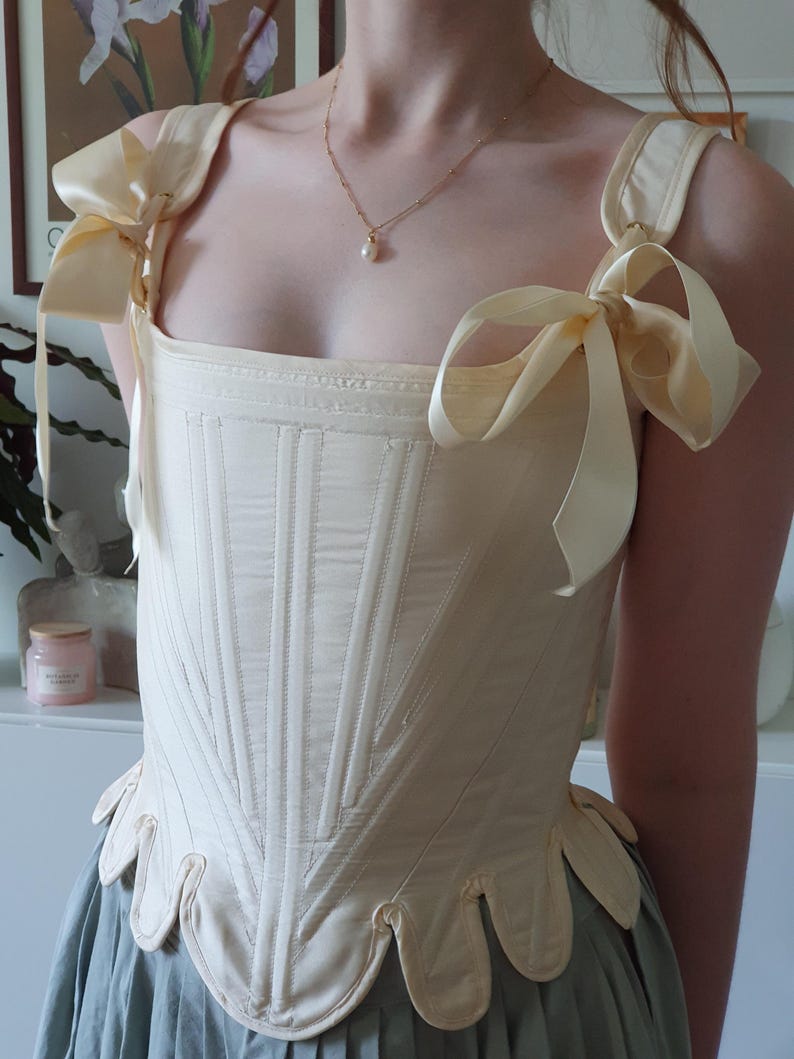 May include: Cream-colored corset with vertical stitching and decorative scalloped hem. The corset features wide shoulder straps tied with large cream-colored bows. A gold necklace with a pearl pendant is also visible.