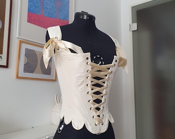 May include: Cream-colored corset with a lace-up back, displayed on a black dress form. The corset features delicate shoulder straps tied with satin bows. The bottom edge has a scalloped design.