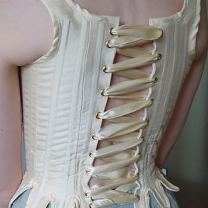 May include: Cream-colored corset with vertical stitching and a laced back. The corset features a decorative, scalloped lower edge and gold-toned eyelets. The back is laced with a cream-colored satin ribbon.