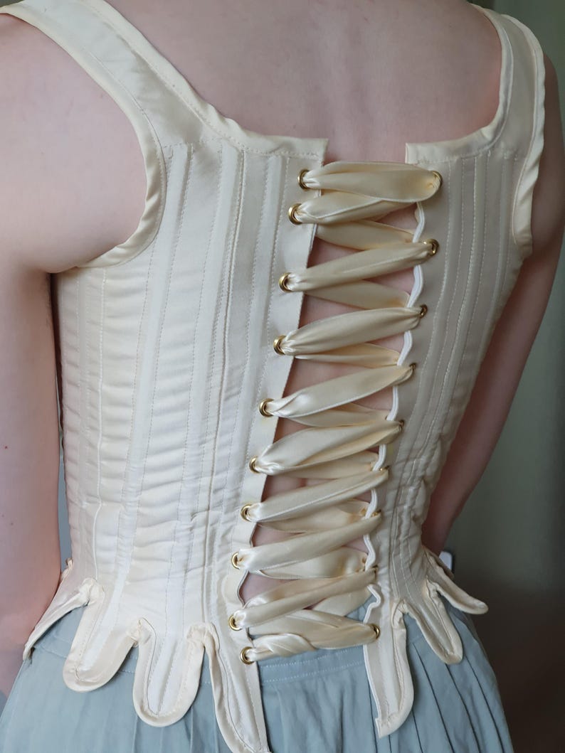 May include: Cream-colored corset with vertical stitching and a laced back. The corset features a decorative, scalloped lower edge and gold-toned eyelets. The back is laced with a cream-colored satin ribbon.