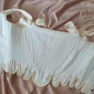May include: Ivory-colored corset with a scalloped hem and ribbon ties. The corset features a structured design with vertical seams and a decorative trim along the bottom edge. The straps are secured with delicate bows, adding a touch of elegance to the garment.