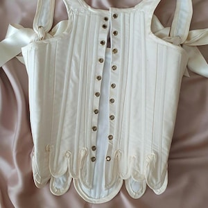 May include: Ivory-colored corset with vertical stitching and a scalloped hem. The corset features shoulder straps tied with satin ribbons and a series of metal eyelets down the front. A classic piece of historical clothing.