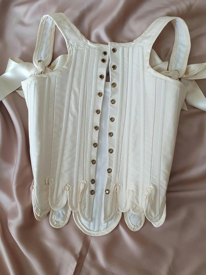 May include: Ivory-colored corset with vertical stitching and a scalloped hem. The corset features shoulder straps tied with satin ribbons and a series of metal eyelets down the front. A classic piece of historical clothing.