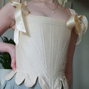 May include: Cream-colored corset with vertical stitching and a scalloped hem. The corset features satin ribbon shoulder straps tied into bows. A vintage-inspired garment, it is likely for historical reenactment or costume.
