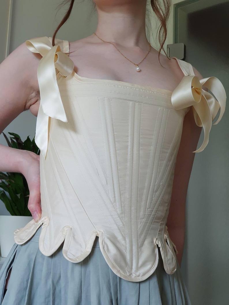 May include: Cream-colored corset with vertical stitching and a scalloped hem. The corset features satin ribbon shoulder straps tied into bows. A vintage-inspired garment, it is likely for historical reenactment or costume.