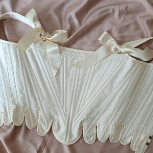May include: Ivory-colored corset with vertical paneling and scalloped edges. The corset features cream-colored ribbon ties at the shoulders and small, decorative buttons. The garment is displayed on a pink satin surface.