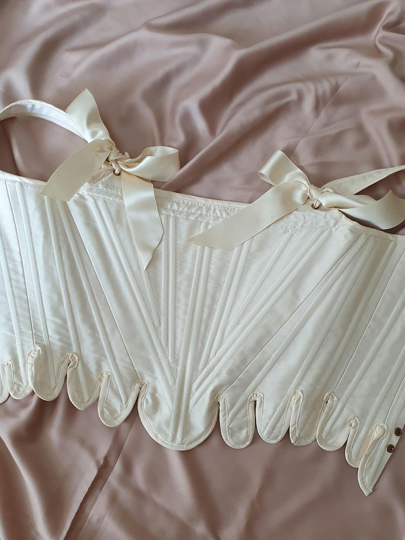 May include: Ivory-colored corset with vertical paneling and scalloped edges. The corset features cream-colored ribbon ties at the shoulders and small, decorative buttons. The garment is displayed on a pink satin surface.