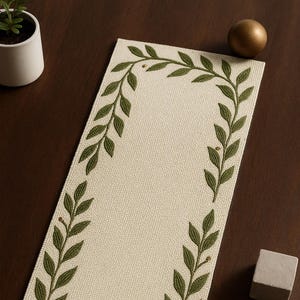Pode incluir: Caminho de mesa retangular creme com uma borda de design de folhas verdes. As folhas são bordadas ao redor das bordas do caminho de mesa. O caminho de mesa está sobre uma superfície marrom escuro. Um pequeno vaso branco, uma esfera dourada, um pequeno cubo cinza e um castiçal também são visíveis.
