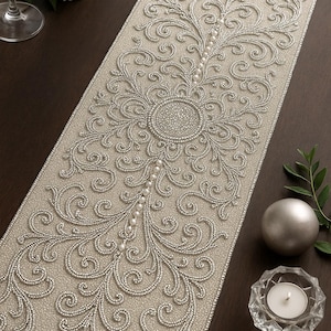 Może przedstawiać: Beżowy bieżnik na stół ze skomplikowanym, koralikowym wzorem kwiatowym. Bieżnik posiada centralny koralikowy medalion i koralikowe akcenty. Perłowa koralikowa obwódka obramowuje krawędzie. Prezentowany na ciemnobrązowej powierzchni, z elementami dekoracyjnymi.