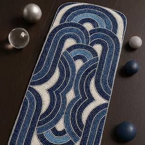 Op de afbeelding: Een kralen loper met een herhalend golfpatroon in blauw- en wittinten. De loper is rechthoekig en heeft een textuur. Decoratieve bollen en een kandelaar zijn rond de loper geplaatst.