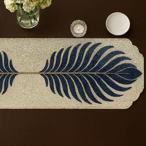 May include: A beaded table runner with a cream-colored background and a symmetrical design of dark blue feather-like shapes. The runner is rectangular with a scalloped edge. Other items on the table include a blue glass, a candle, a white plate, and a vase of flowers.