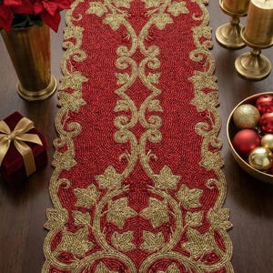 Może przedstawiać: Czerwony, koralikowy bieżnik na stół ze złotymi wzorami liści i winorośli. Prostokątny bieżnik ma szczegółowy wzór. Na zdjęciu znajdują się również złote świece, pudełko na prezent i ozdoby świąteczne.