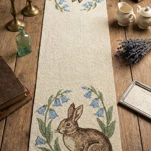 Könnte beinhalten: Beiger Perlen-Tischläufer mit gestickten braunen Hasen und Glockenblumen-Blütenmustern. Der Läufer zeigt einen sitzenden Hasen unten und einen kopfüber hängenden Hasen oben. Dekorative Wohnkultur.