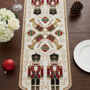 May include: A beaded Christmas table runner with a white background and gold trim. The runner features repeating patterns of nutcrackers, drums, and trumpets in red, green, black, and gold. The runner is approximately 60 inches long.