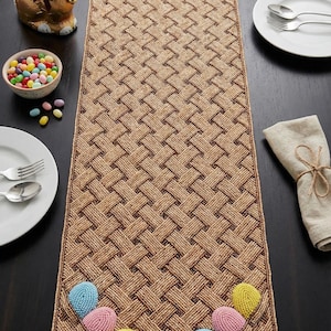 Op de afbeelding: Een bruine, geweven paas tafelloper met kleurrijke, kralen paaseieren. De loper ligt op een donkere houten tafel, met borden, bestek en een chocolade paashaas. Een kom met jelly beans is ook aanwezig.