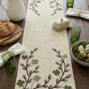 May include: Cream-coloured table runner featuring embroidered branches and green leaves. The runner is decorated with a floral design and is placed on a wooden table. Other items include a white jug, bread, and green glasses.