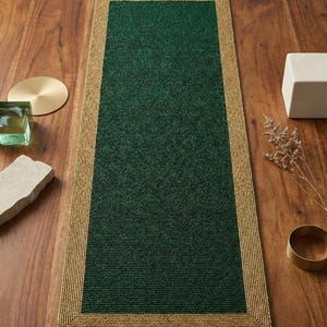 Può includere: Un tappeto rettangolare verde con un bordo dorato bordato di perline. Il tappeto è centrato su una superficie di legno, con oggetti decorativi come un sottobicchiere in ottone, un cubo bianco e un piccolo contenitore di vetro.