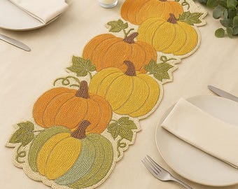 May include: A beaded pumpkin table runner in shades of orange, yellow, and green. The runner features multiple pumpkin shapes with green leaf accents. The runner is placed on a beige tablecloth, with plates, silverware, and a candle in the background.