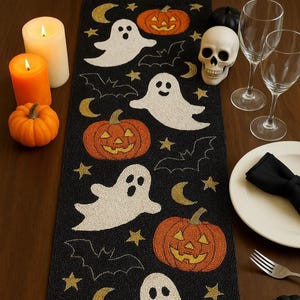 May include: A black Halloween table runner with beaded white ghosts, orange pumpkins, gold stars, and crescent moons. Lit candles and a small pumpkin are on the left. Wine glasses and a skull are on the right.