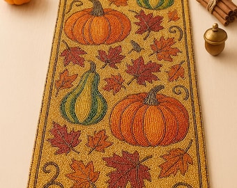 May include: A beaded table runner with an autumn theme. The runner features orange pumpkins, green gourds, and red and orange fall leaves on a gold background. Decorative elements include a small lantern, cinnamon sticks, and an acorn.