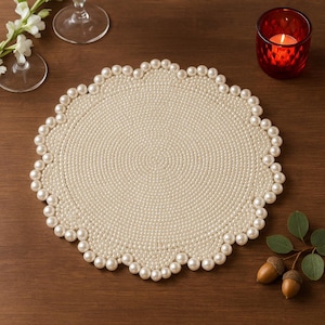 May include: Cream-coloured round placemat with a scalloped edge decorated with faux pearls. The placemat has a textured surface and is set on a dark wooden table. A red candle holder with a lit candle is visible.