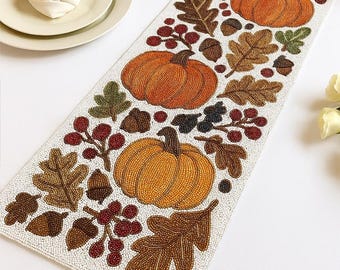 May include: Beaded table runner featuring pumpkins, oak leaves, acorns, and berries in shades of orange, brown, and red. The runner is white with a beaded texture, perfect for fall or Thanksgiving decor.