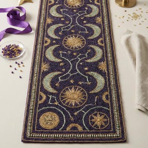 May include: A dark purple beaded table runner with celestial designs. The runner features gold suns, moons, and stars, with a border of small beads. The design is symmetrical and detailed, creating an elegant look.