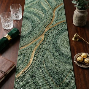 May include: A beaded green and gold table runner with a flowing, organic design. The runner is rectangular and features a pattern of swirling lines in shades of green and gold. The runner is on a dark wooden table, with glasses, a napkin, and other decorative items.