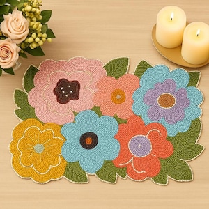 May include: A beaded floral table runner featuring a colorful array of flowers in pink, orange, blue, yellow, and purple. The flowers have brown, orange, and purple centers and are surrounded by green leaves. The runner is approximately 12 inches wide and 24 inches long.