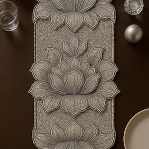 May include: A beaded, silver-colored table runner with three large lotus flower designs. The runner is rectangular with rounded edges and is placed on a dark brown surface. The flowers are detailed with small beads.