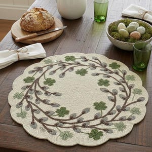 May include: Cream-coloured beaded placemat with a scalloped edge, featuring a circular design of brown branches and green shamrocks. The placemat is on a dark wooden table, alongside a loaf of bread, a jug, and green glasses.