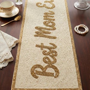 Puede incluir: Camino de mesa de cuentas color crema con las palabras "Best Mom Ever" en escritura dorada. El camino de mesa tiene un borde dorado con cuentas. La imagen también muestra una taza de té, flores y una pulsera de perlas.