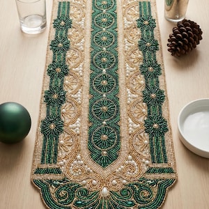 May include: An ornate, beaded table runner in shades of green and gold. The runner features a central design with repeating floral and geometric patterns, accented with small white beads. The edges are scalloped and detailed, creating an elegant, decorative piece.