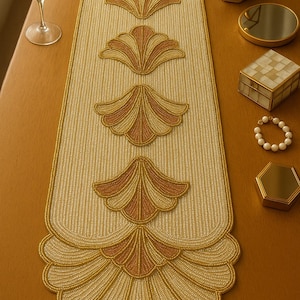 Może przedstawiać: Beżowo-złoty, koralikowy bieżnik na stół z powtarzającym się wzorem w kształcie wachlarza. Bieżnik zdobiony jest misternymi koralikami i ma falisty brzeg. Na stole znajdują się również świeca, lustro i szkatułka na biżuterię. Długość bieżnika to około 150 cm.