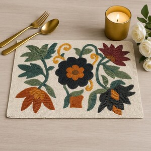May include: Beige beaded placemat with a floral design in shades of orange, green, and black. Gold-colored fork and spoon are to the left. A lit candle in a gold holder and white roses are to the right.