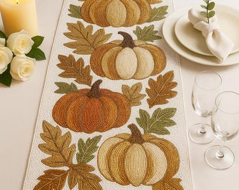 May include: Beaded table runner featuring a repeating pattern of pumpkins and fall leaves in shades of orange, gold, and green. The runner is on a white background, with a candle, flowers, and place settings visible.