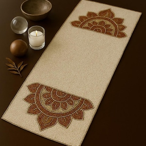 Op de afbeelding: Beige rechthoekige tafelloper met een getextureerd oppervlak en decoratieve bloemmotieven in bruin- en goudtinten aan beide uiteinden. De loper ligt op een donkerbruin oppervlak, wat een contrast creëert. Een kaars, kom en decoratieve bollen staan op de achtergrond.