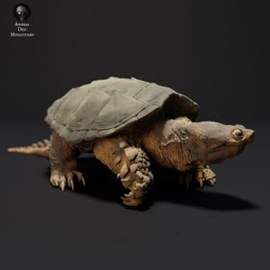 May include: A detailed miniature of a snapping turtle, featuring a textured, gray-green shell and a tan body. The sculpture has a realistic appearance, with intricate details on the head, legs, and tail. The Animal Den Miniatures logo is in the upper left corner.