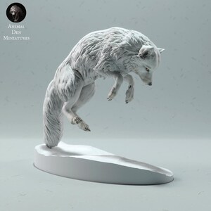 Op de afbeelding: Een witte sculptuur van een springende poolvos met gedetailleerde vachttextuur en amberkleurige ogen. De vos poseert in de lucht boven een met sneeuw bedekte basis. Van Animal Den Miniatures.