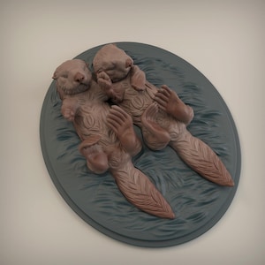Può includere: Scultura di due lontre sdraiate sulla schiena, di colore marrone chiaro. Le lontre sono posizionate su una base ovale grigia con una superficie strutturata. Le lontre hanno una pelliccia dettagliata e sono in posa rilassata.