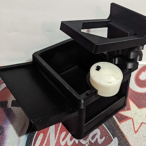 May include: A black 3D-printed device with a rectangular base, a sliding tray, and an adjustable top section. A white cylindrical component is visible within the device. The device appears to be a tool or accessory.