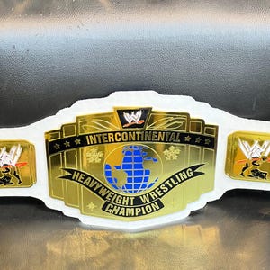 May include: A gold-colored WWE Intercontinental Heavyweight Wrestling Champion belt featuring a central globe graphic in blue, black text on gold plates, and a white strap. The plates display the WWE logo and the text Intercontinental Heavyweight Wrestling Champion.