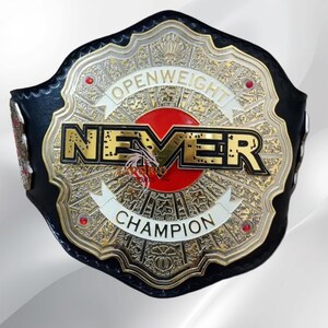 Könnte beinhalten: Ein schwarzer Meisterschaftsgürtel mit einer großen, verzierten Mittelplatte aus gold- und silberfarbenem Metall. Die Platte zeigt den Text "OPENWEIGHT NEVER CHAMPION" in fetter Schrift, zentriert über einem roten runden Emblem mit vier kleinen roten Steinen.