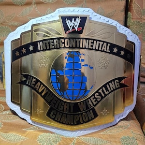 May include: A WWE Intercontinental Heavyweight Wrestling Champion belt featuring a large gold-colored central plate with a blue globe graphic, black accents, and the WWE logo at the top, all mounted on a white leather-like strap with scalloped edges.