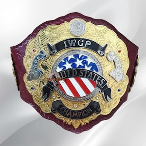 Könnte beinhalten: Ein Wrestling-Meisterschaftsgürtel mit einem dunkelroten Lederriemen und einer großen, verzierten goldfarbenen Metallplatte. Die Platte zeigt den Text IWGP, UNITED STATES und CHAMPION sowie ein zentrales kreisförmiges Emblem der US-Flagge.