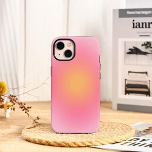 Op de afbeelding: Telefoonhoesje met roze en gele gradiënt. De hoes heeft een gladde afwerking en een zwarte cameramodule. Het ontwerp heeft een zachte gradiënt van roze naar geel in het midden. De telefoonhoes ligt op een geweven onderzetter.