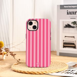 May include: Pink and light pink striped phone case. The case has a black border and a camera cutout. The initials "CE" are at the bottom of the case. The phone case is on a round, woven coaster.