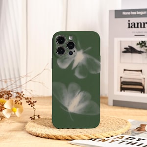 Puede incluir: Funda de teléfono verde oscuro con un diseño de mariposa blanca. La funda tiene un acabado mate y un recorte para la cámara. El diseño se repite en la funda. La funda del teléfono está sobre un posavasos tejido.