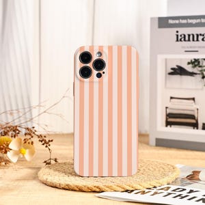 Könnte beinhalten: Eine Handyhülle mit Pfirsich- und Weißstreifen. Die Hülle hat ein vertikales Streifenmuster und ein Kameramodul mit mehreren Linsen. Die Handyhülle liegt auf einem gewebten Untersetzer, mit einem Buch und dekorativen Elementen im Hintergrund.